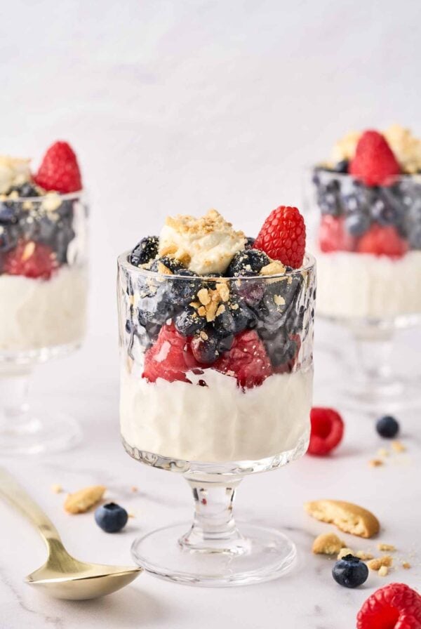 Three cheesecake parfaits, each topped with blueberries, raspberries, and crumbled vanilla cookies in footed parfait glasses, surrounded by cookie crumbs, whole berries, and a spoon.