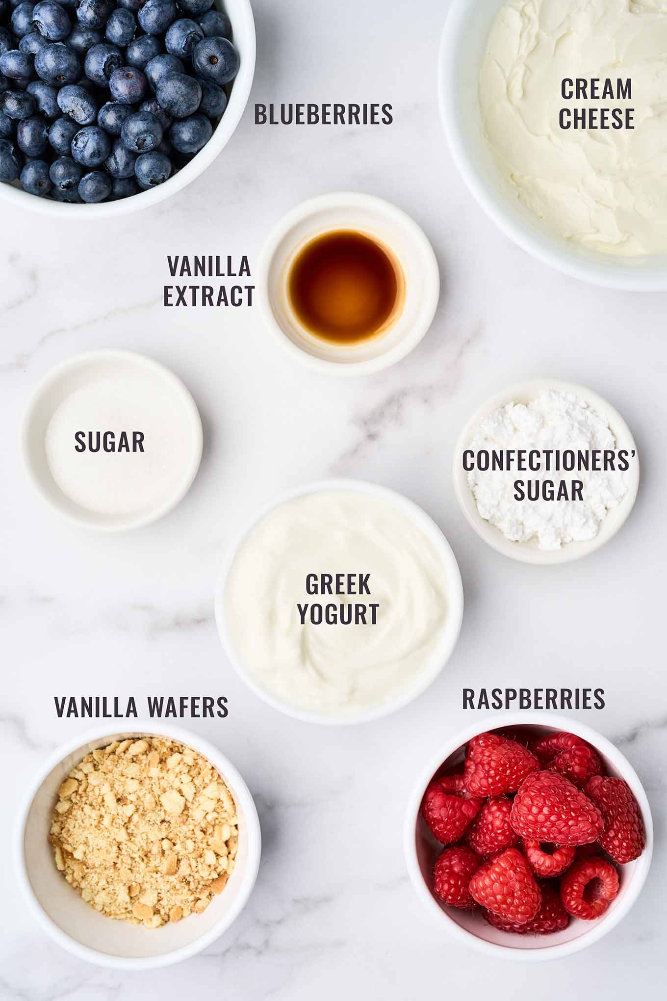 An overhead view of the ingredients for no-bake cheesecake parfaits with fresh berries; cream cheese, Greek vanilla yogurt, confectioners' sugar, vanilla wafers, raspberries, blueberries, and vanilla extract.