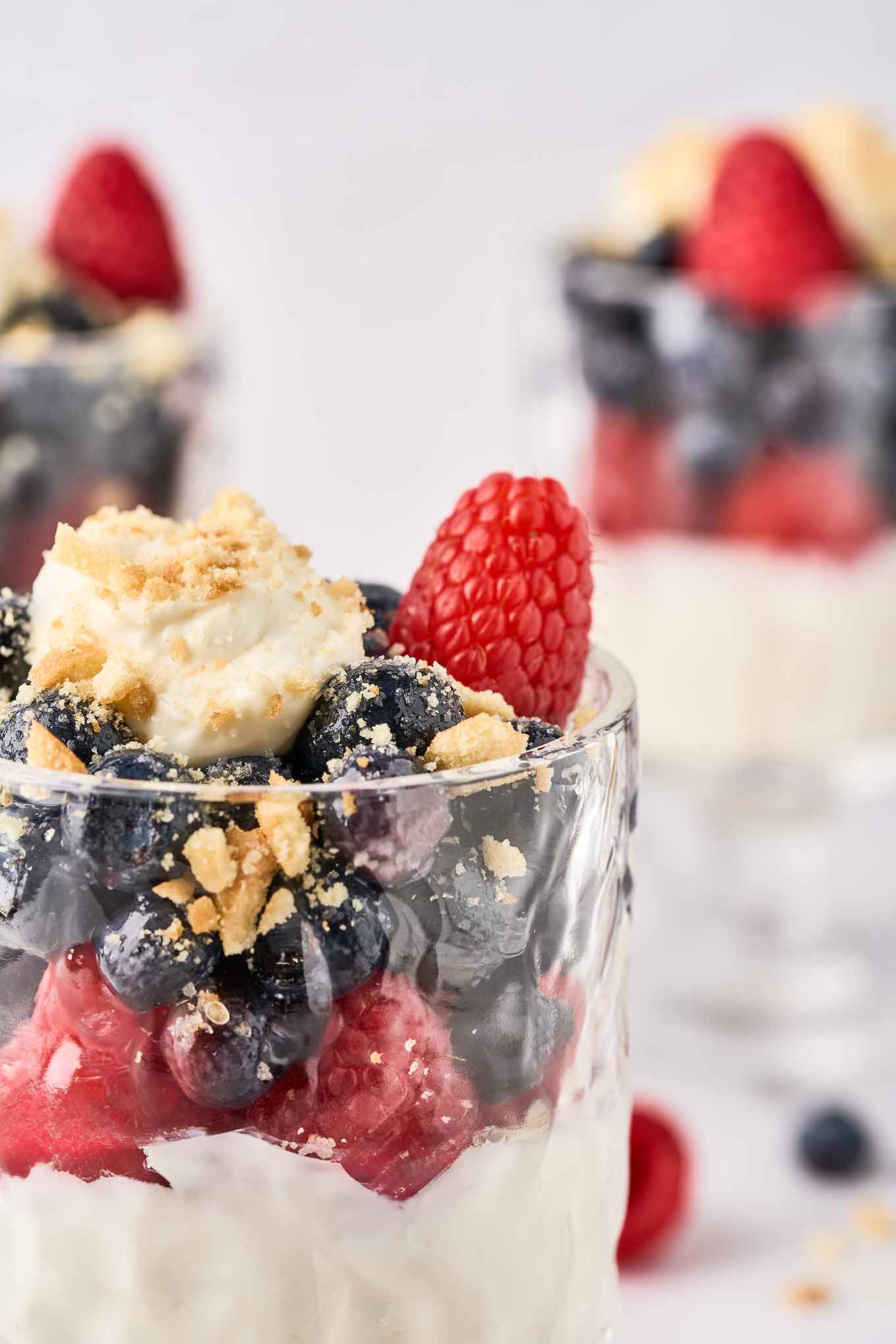 A close-up view of a parfait glass with no-bake cheesecake topped with raspberries, sugared blueberries and crumbled vanilla wafers.