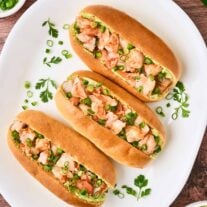 A white oval platter with three split top hot dog buns filled with sautéed shrimp, avocado-cilantro mayo, and chopped scallions, prep bowls with more mayo, chopped jalapeno, and chopped scallions in the background.