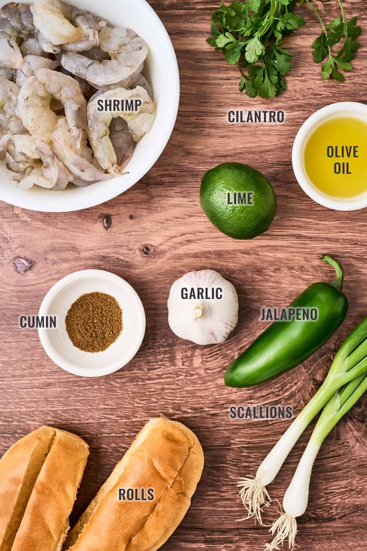 An overhead, labeled view of the ingredients needed to make shrimp rolls with avocado mayonnaise; extra-large shrimp, cilantro, lime, olive oil, garlic, jalapeno, scallions, cumin, and rolls.