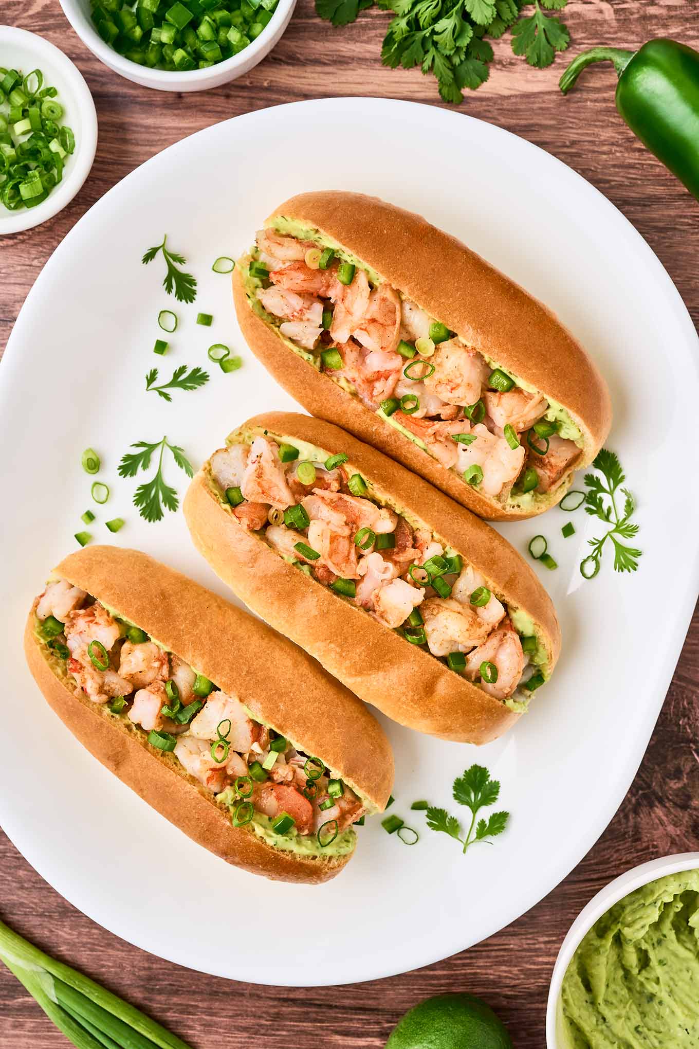 A white oval platter with three split top hot dog buns filled with sautéed shrimp, avocado-cilantro mayo, and chopped scallions, prep bowls with more mayo, chopped jalapeno, and chopped scallions in the background.