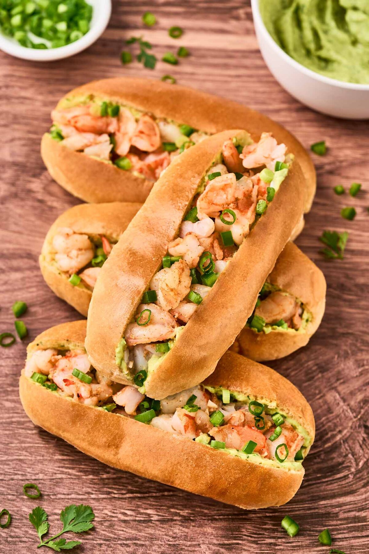 Four shrimp rolls with avocado mayonnaise garnished with chopped scallions arranged on a wooden surface with extra mayo and scallions in bowls in the background.