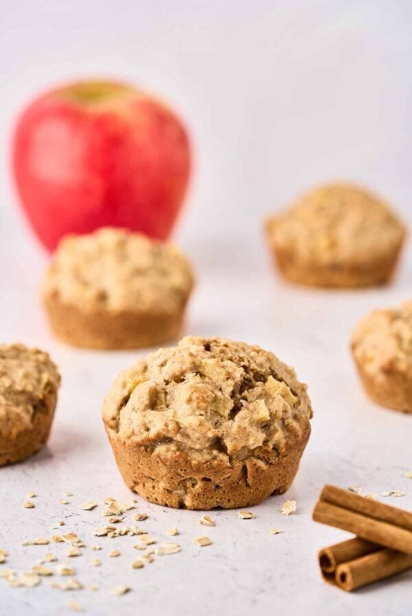 A single apple oatmeal muffin with more muffins and a whole apple in the background, cinnamon sticks and scattered oats in the foreground.
