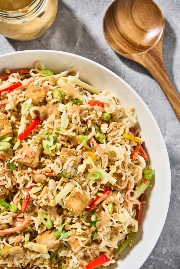 A serving bowl of Asian noodle salad with peanut dressing and crispy, golden tofu, extra peanut dressing and salad servers to the side.