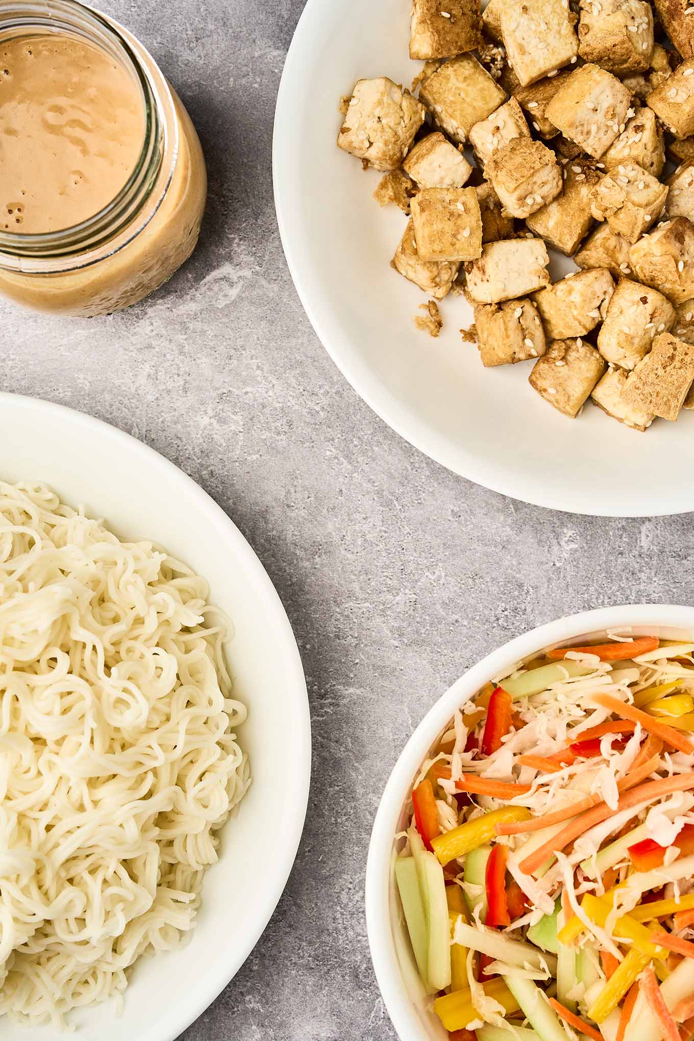 Asian noodle salad ingredients ready to be assembled; a bowl of slivered bell peppers and cucumbers combined with shredded carrots and cabbage, another bowl of cooked ramen noodles, a bowl of crispy tofu, and a jar of peanut dressing.