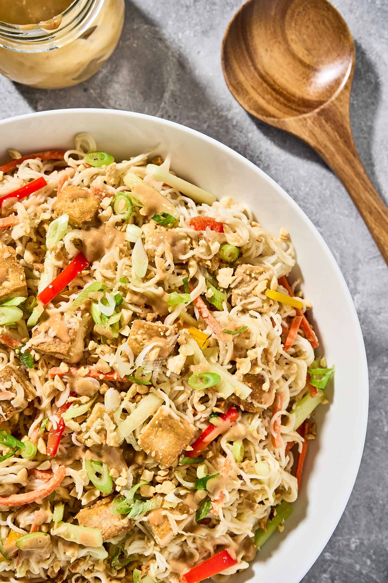 A serving bowl of Asian noodle salad with peanut dressing and crispy, golden tofu, extra peanut dressing and salad servers to the side.