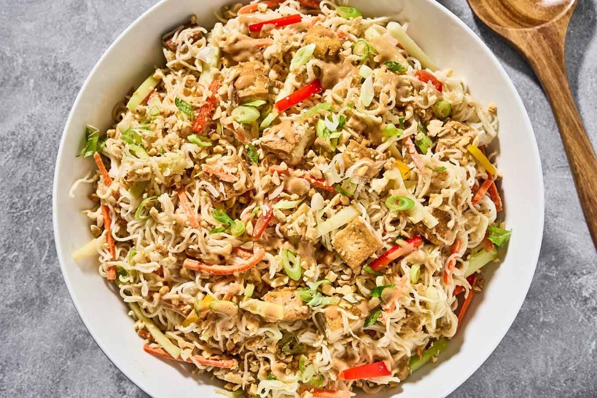 A serving bowl filled with cold Asian noodle salad tossed with peanut dressing. Ramen noodles, crispy golden tofu, cabbage, bell pepper, cucumber, carrots, scallions, crushed peanuts, and sesame seeds.