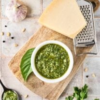 A bowl of basil and parsley pesto, fresh basil leaf, cheese grater, and a wedge of parmesan cheese on a wooden cutting board, surrounded by fresh parsley and basil, and other pesto sauce ingredients.