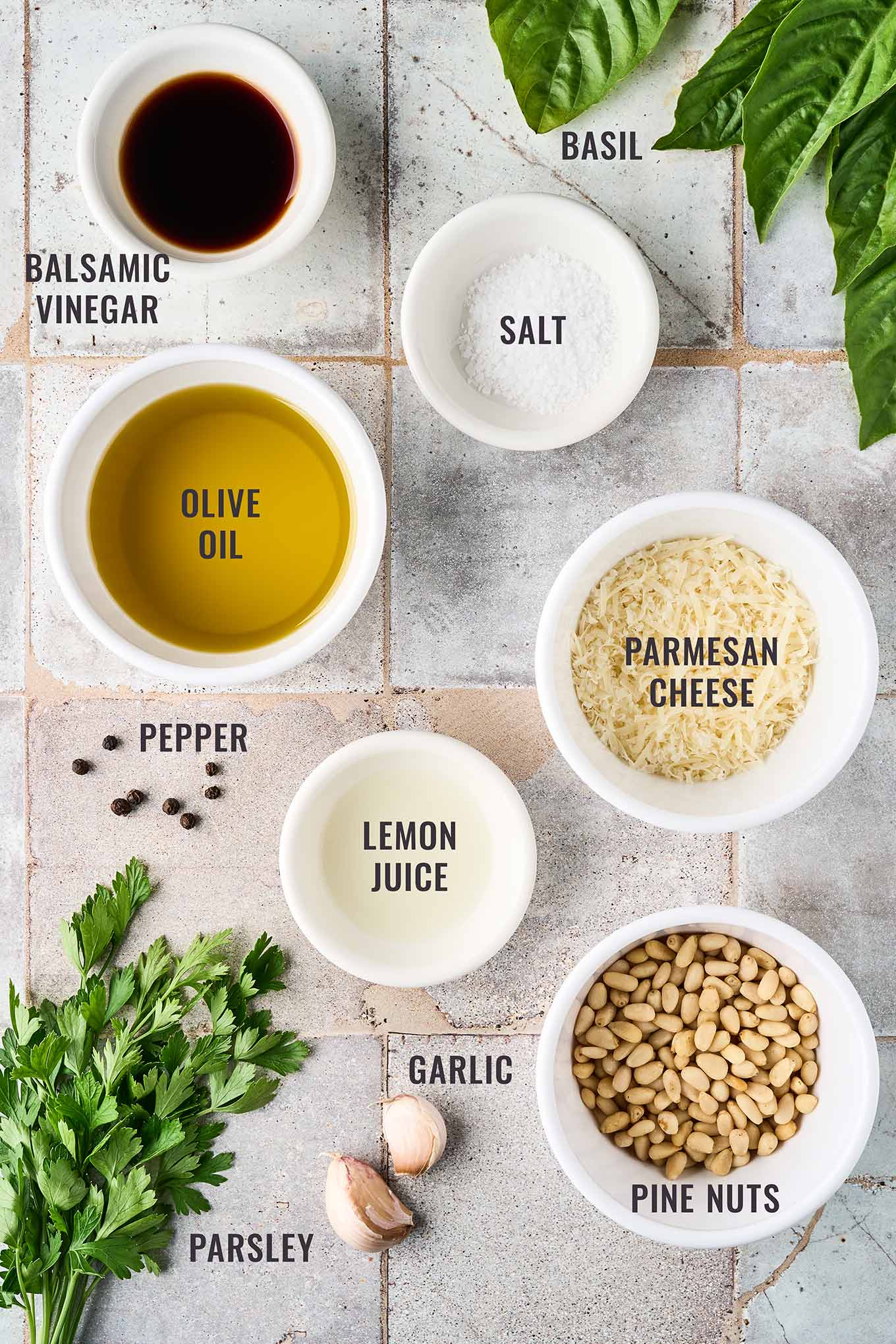 A labeled, overhead view of the ingredients for basil and parsley pesto; fresh basil, parsley, pine nuts, Parmesan cheese, garlic, lemon juice, pepper, olive oil, salt, and balsamic vinegar.