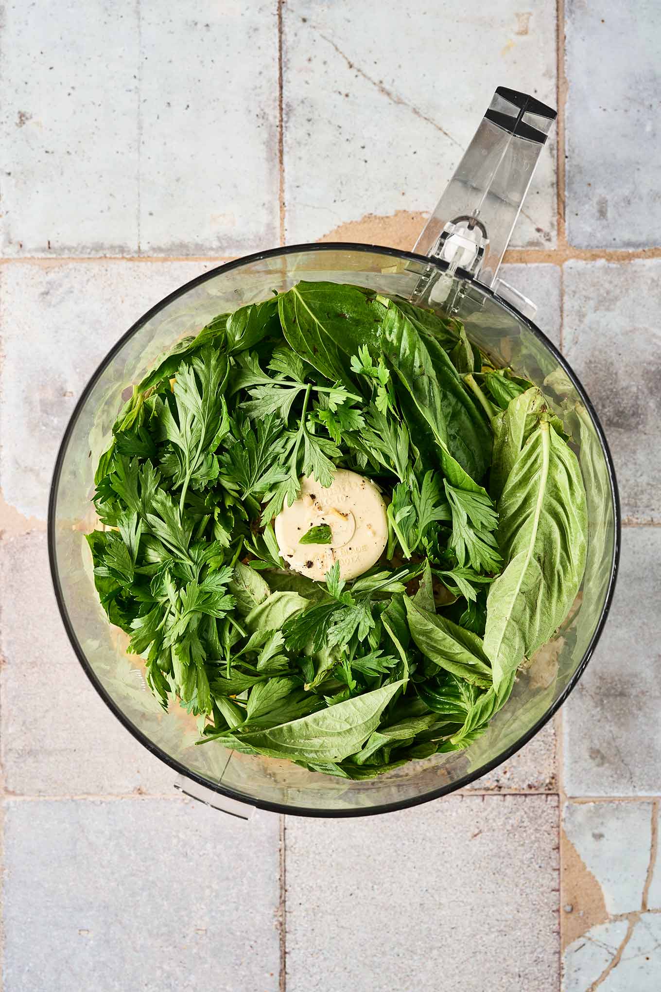 An overhead view of the ingredients for basil and parsley pesto in a food processor, ready to be blended.