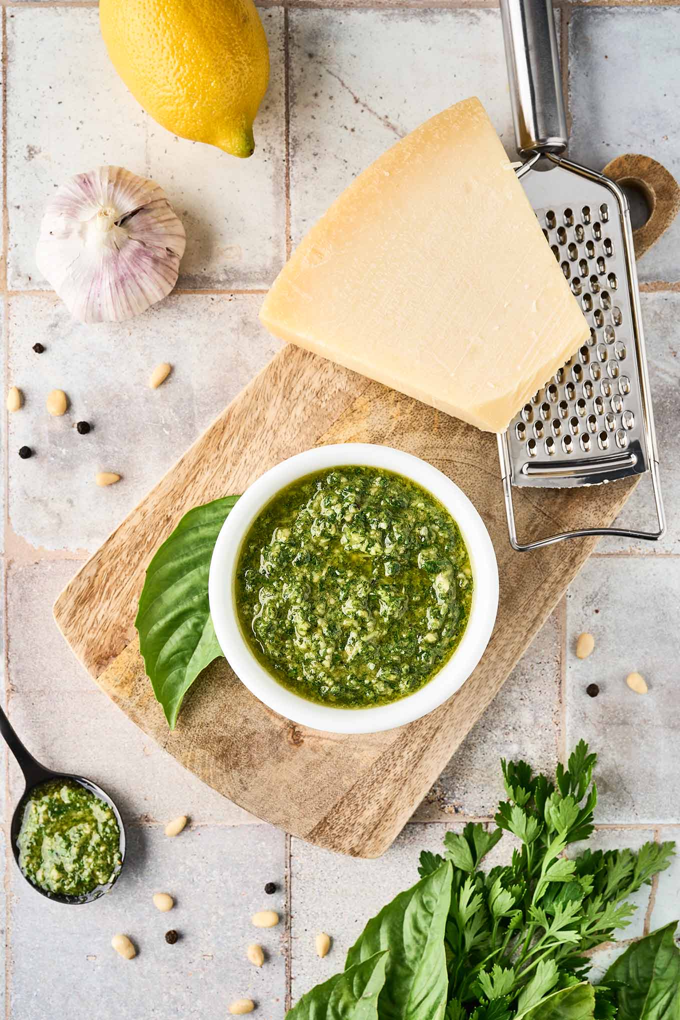 A bowl of basil and parsley pesto, fresh basil leaf, cheese grater, and a wedge of parmesan cheese on a wooden cutting board, surrounded by fresh parsley and basil, and other pesto sauce ingredients.