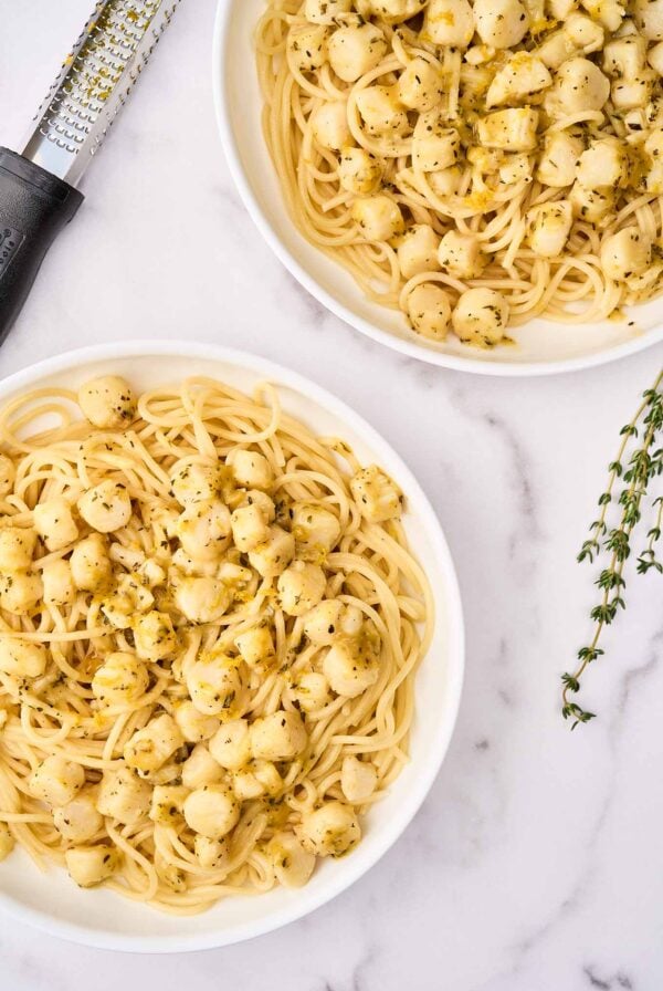 Two servings of bay scallop pasta with Sicilian citrus sauce in shallow white bowls, surrounded by a sprig of fresh thyme, and a citrus zester.