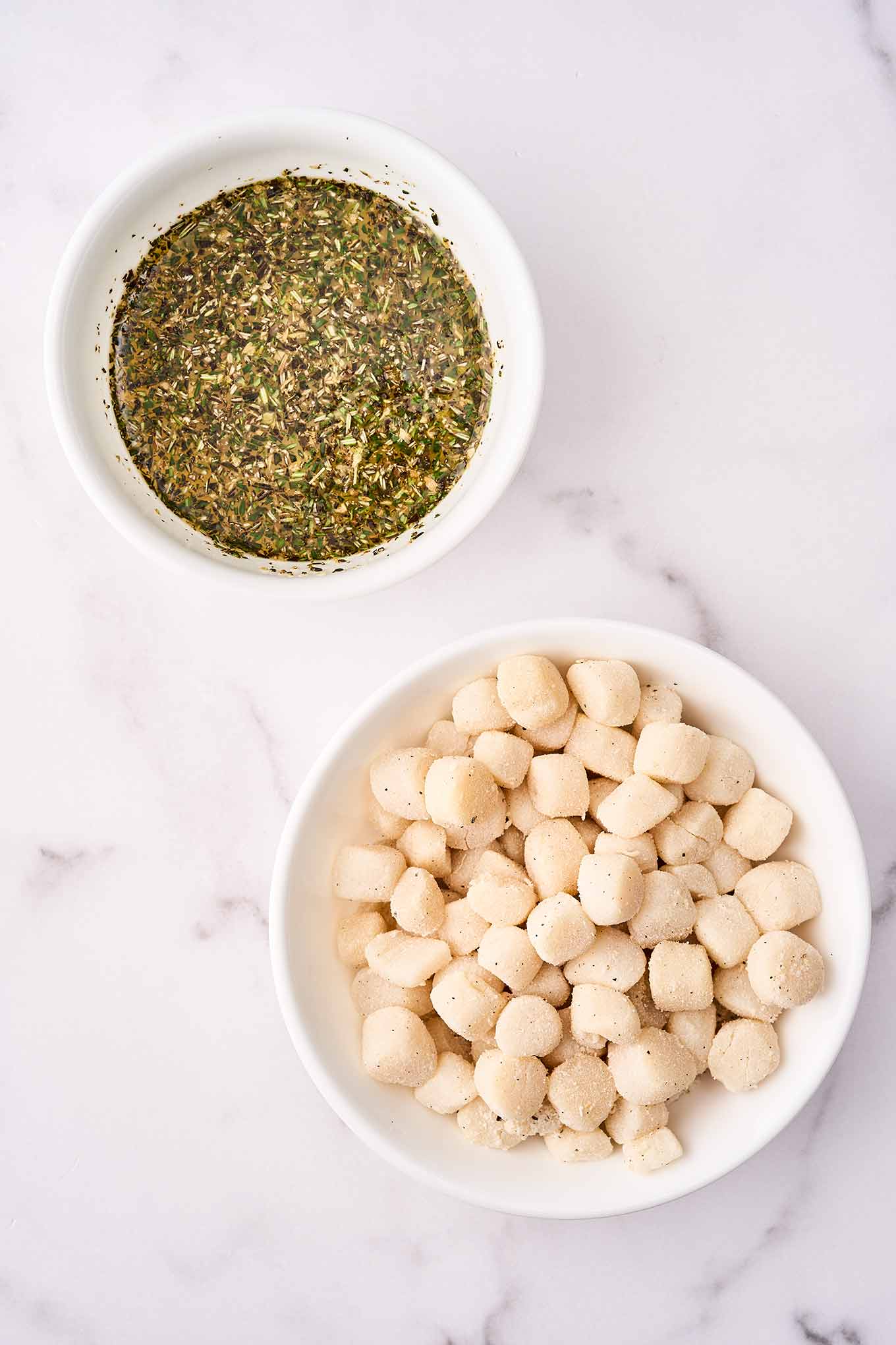 Two small mixing bowls, one filled with bay scallops dusted in seasoned flour, the other with fresh thyme, rosemary, and oregano.