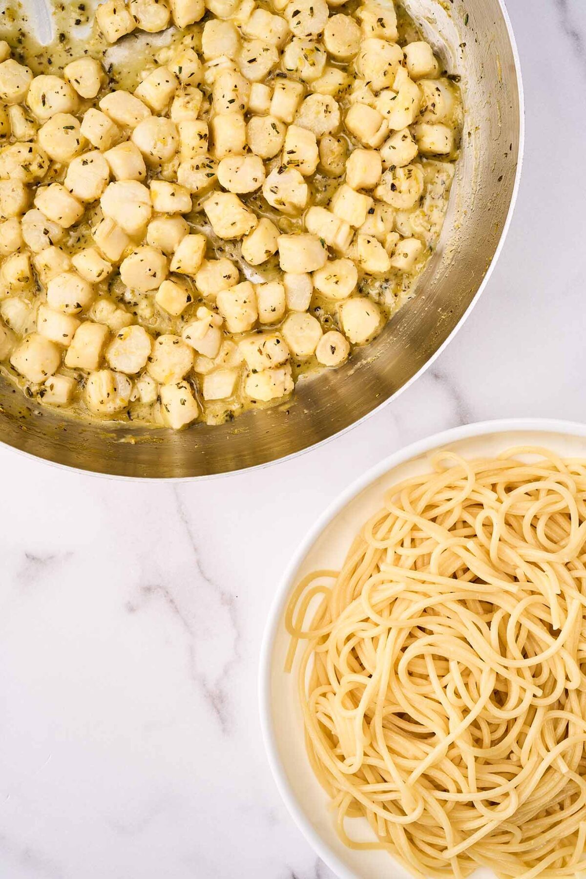 A frying pan with sautéed bay scallops in citrus, herb, and olive oil sauce next to a bowl of cooked thin spaghetti, ready to be combined.