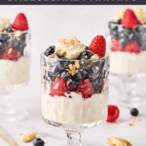 Three cheesecake parfaits, each topped with blueberries, raspberries, and crumbled vanilla cookies in footed parfait glasses, surrounded by cookie crumbs, whole berries, and a spoon.