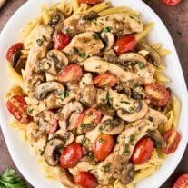 An overhead view of chicken Marsala pasta on a white oval serving platter, surrounded by a scattering of halved grape tomatoes, a sprig of parsley, and a wooden spoon.