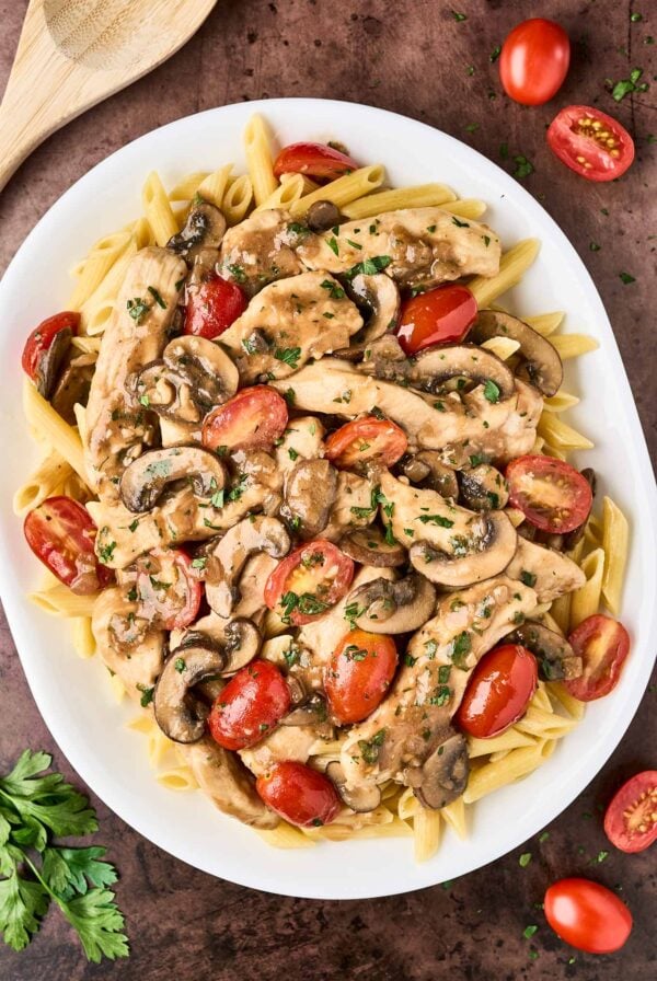 An overhead view of chicken Marsala pasta on a white oval serving platter, surrounded by a scattering of halved grape tomatoes, a sprig of parsley, and a wooden spoon.