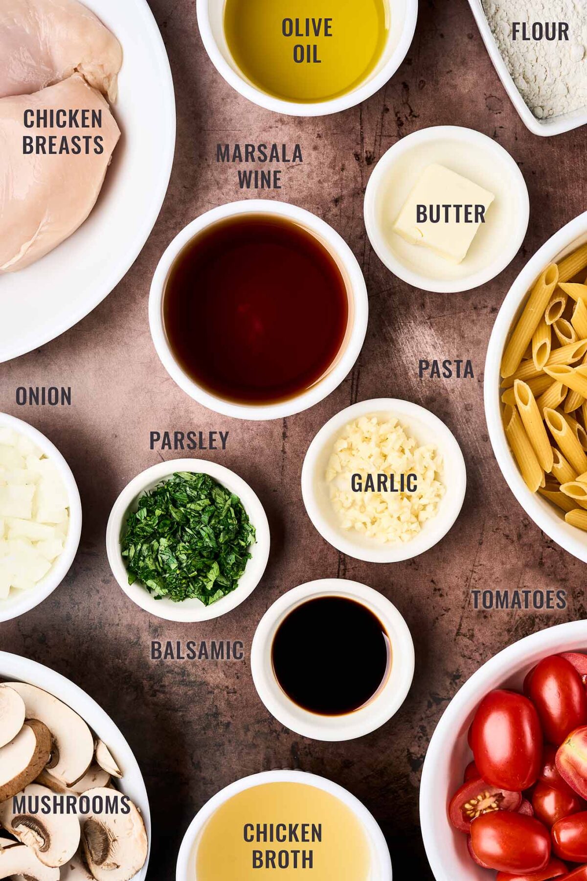 A labeled, overhead view of the ingredients for chicken Marsala pasta; chicken breasts, Marsala wine, penne pasta, mushrooms, garlic, onion, grape tomatoes, and more.