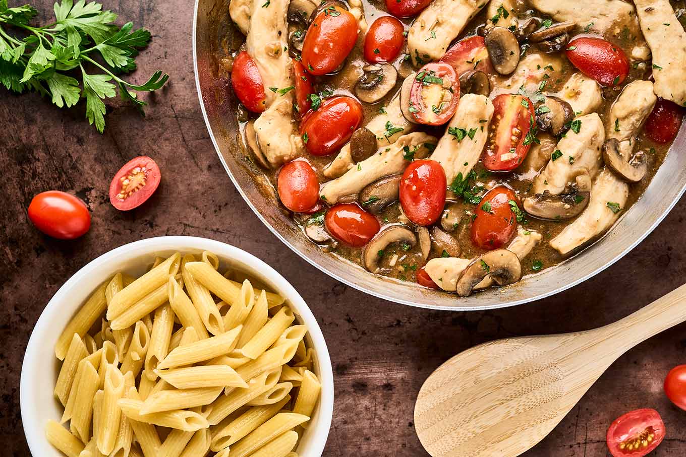 A frying pan filled with chicken Marsala with fresh tomatoes next to a bowl of cooked penne pasta, ready to be combined to finish the dish.