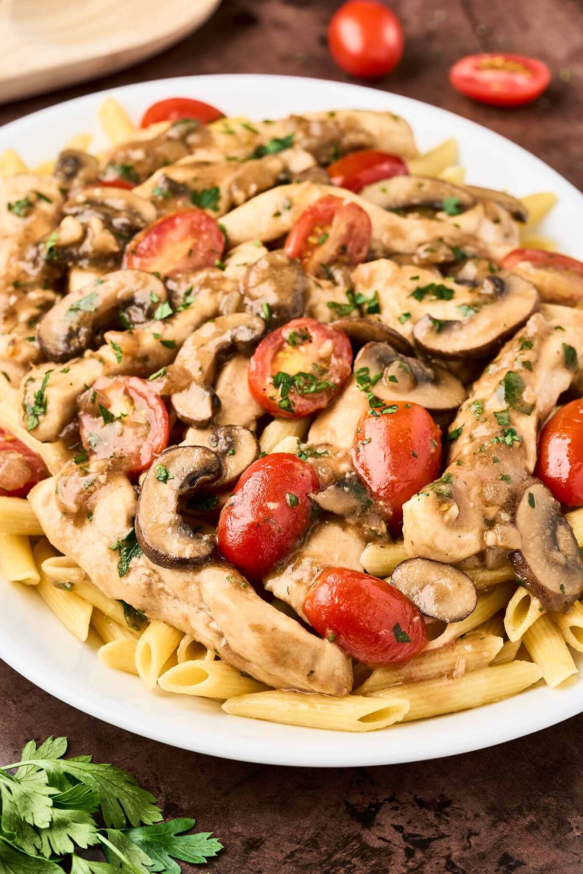 A serving platter of penne pasta topped with chicken Marsala with fresh tomatoes, garnished with chopped parsley.