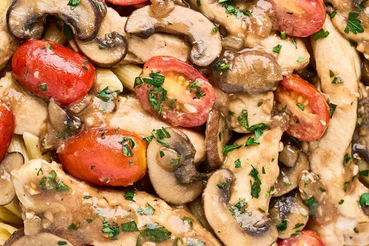 A close up look at chicken Marsala pasta showing chicken breast strips, penne pasta, mushrooms, grape tomatoes, bits of garlic, and chopped parsley.