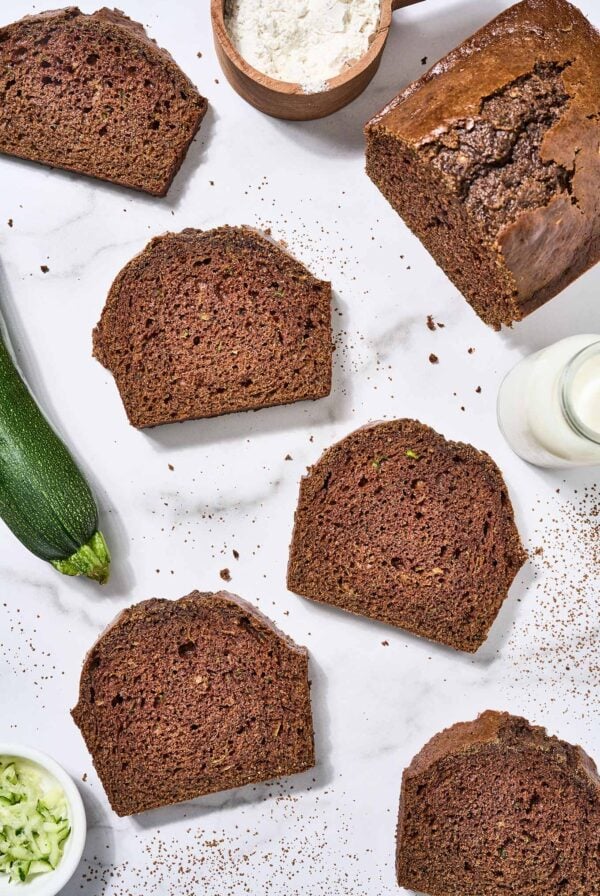A partially sliced loaf of zucchini bread on a marble surface surrounded by slices of bread, a prep bowl of shredded zucchini, a whole zucchini, cup of flour, and pitcher of buttermilk.