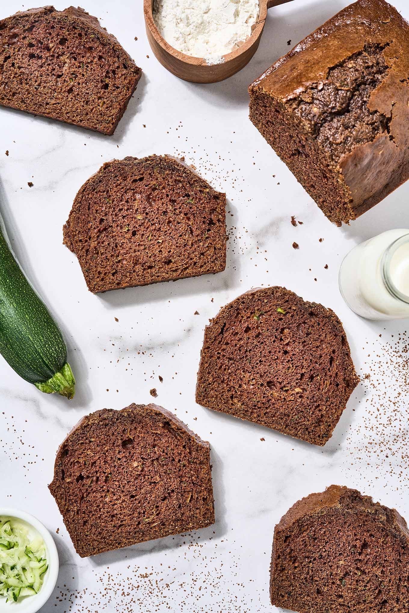 A partially sliced loaf of zucchini bread on a marble surface surrounded by slices of bread, a prep bowl of shredded zucchini, a whole zucchini, cup of flour, and pitcher of buttermilk.