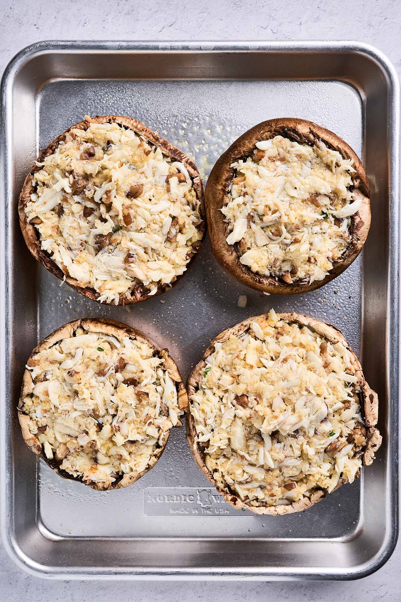 A baking sheet with four portobello mushroom caps filled with a combination of lump crabmeat, panko, and grated Muenster cheese.