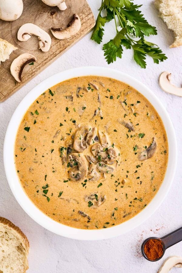 An overhead view of a bowl of creamy Hungarian mushroom soup surrounded by a board with sliced fresh mushrooms, a sprig of parsley, a spoonful of paprika and slices of crust bread for serving.