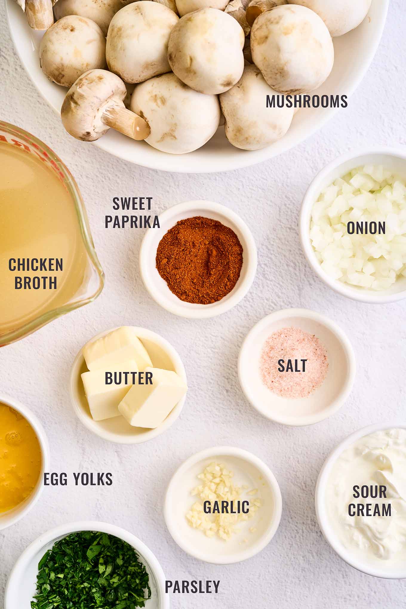 A labeled, overhead view of the ingredients needed to make creamy Hungarian mushroom soup; fresh mushrooms, chicken broth, sweet paprika, onion, garlic, butter, salt, sour cream, egg yolks, and fresh parsley.
