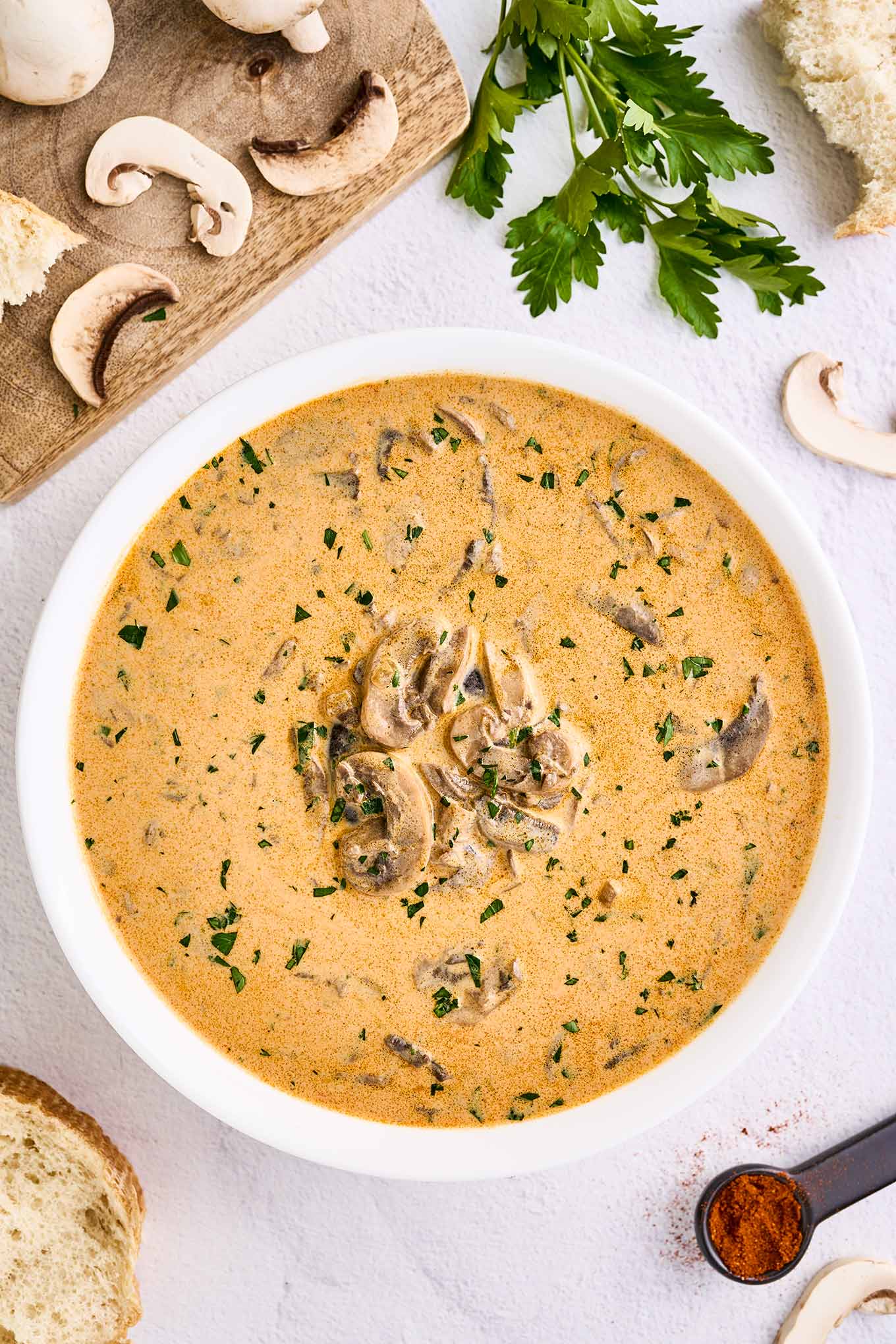 An overhead view of a bowl of creamy Hungarian mushroom soup surrounded by a board with sliced fresh mushrooms, a sprig of parsley, a spoonful of paprika and slices of crust bread for serving.