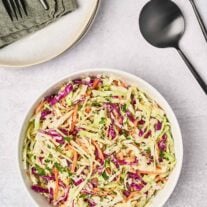 A serving bowl filled with creamy, light coleslaw, black salad servers to the right, and a stack of plates, topped with linen napkins, and utensils to the rear.