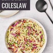 A serving bowl filled with creamy, light coleslaw, black salad servers to the right, and a stack of plates, topped with linen napkins, and utensils to the rear.