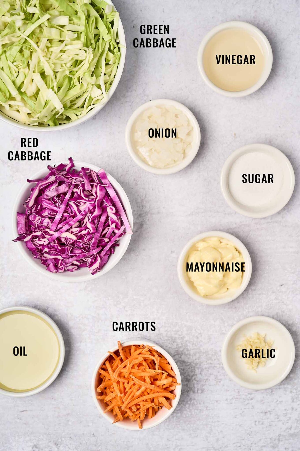 A labeled, overhead view of the ingredients needed for creamy, light, coleslaw; thin sliced red and green cabbage, chopped onion, shredded carrots, garlic, oil, vinegar, and sugar.