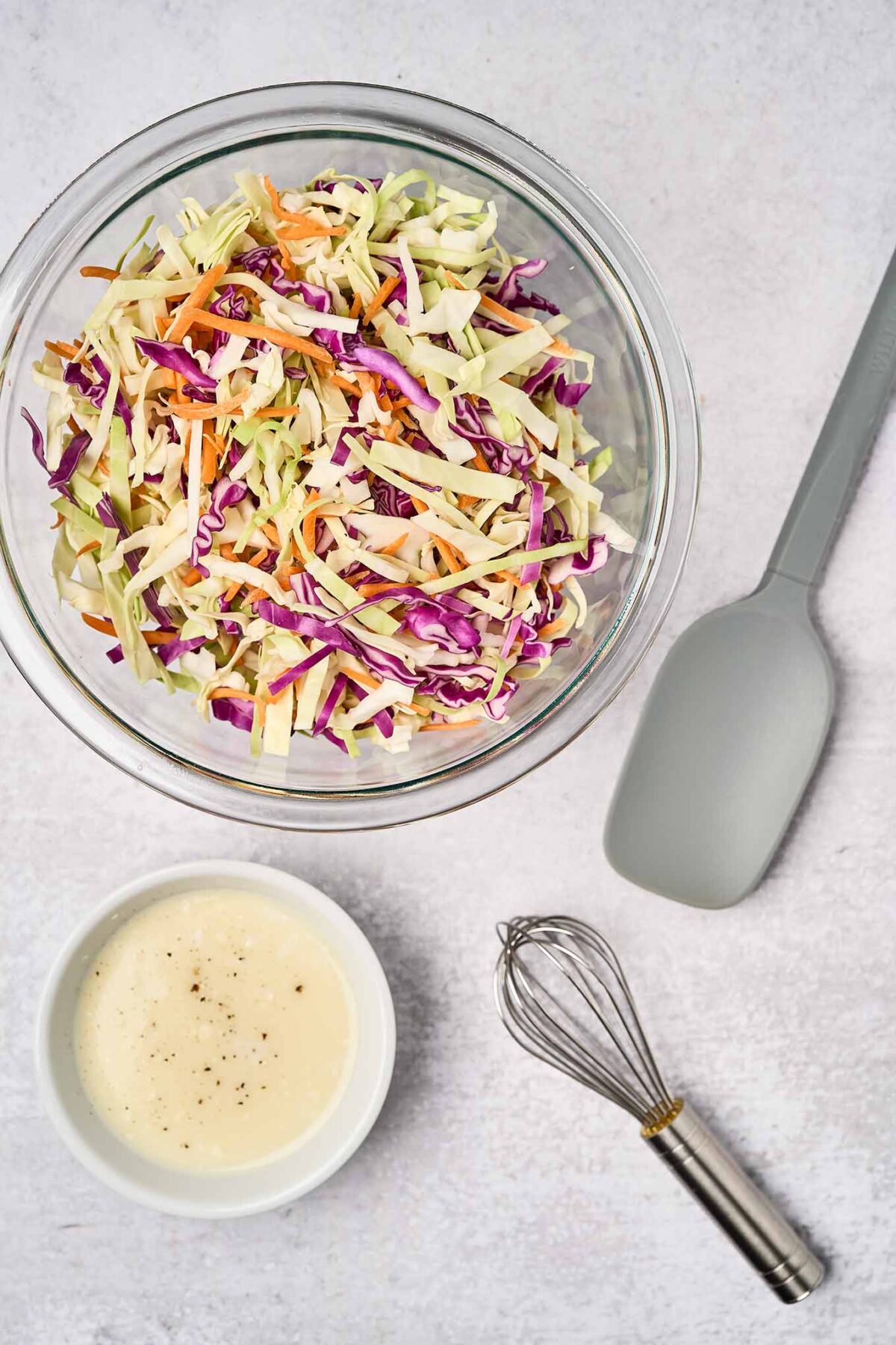 A large mixing bowl filled with shredded cabbage and carrots, and a small mixing bowl filled with light, creamy coleslaw dressing, a silicone spatula and wire whisk to the right.