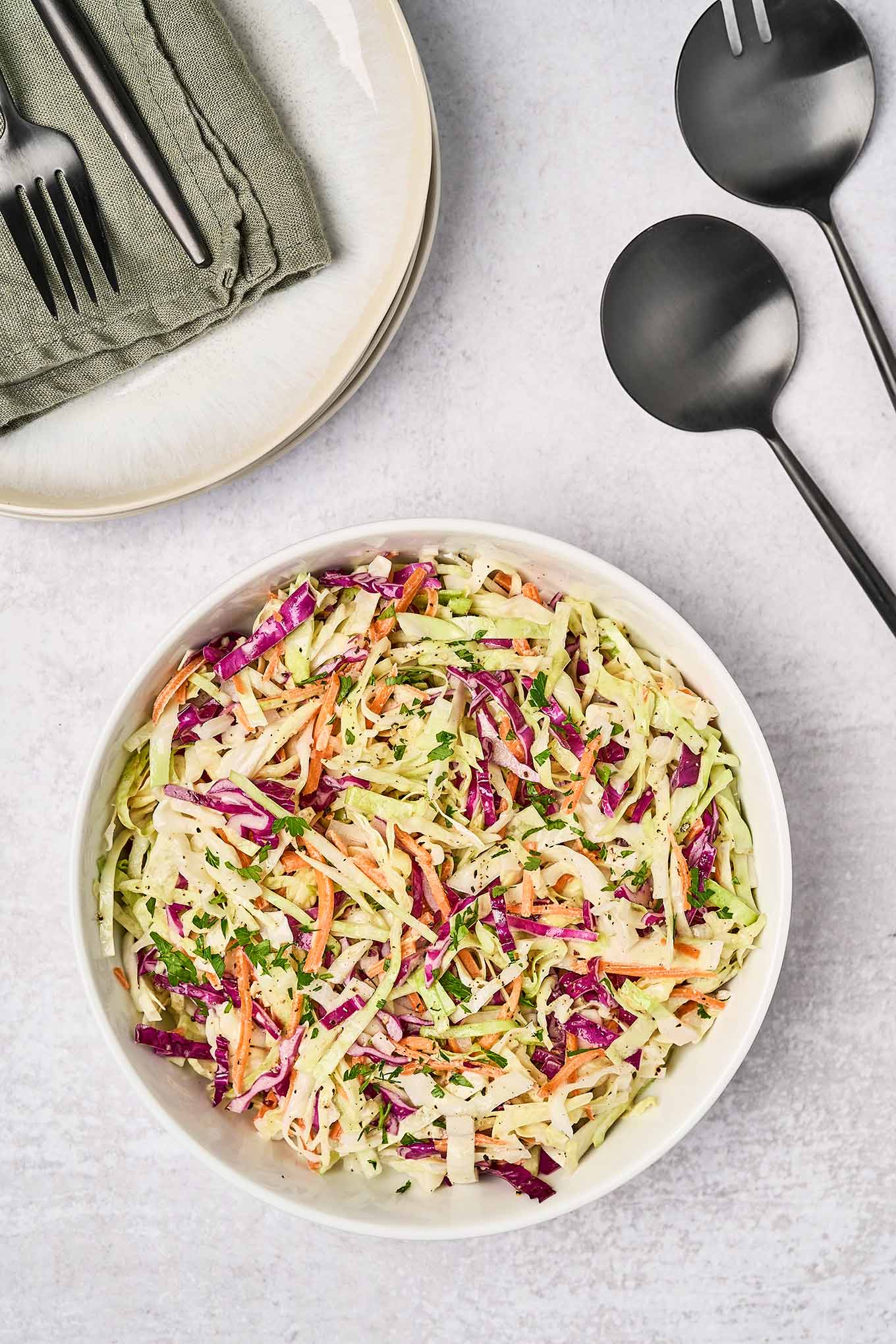 A serving bowl filled with creamy, light coleslaw, black salad servers to the right, and a stack of plates, topped with linen napkins, and utensils to the rear.