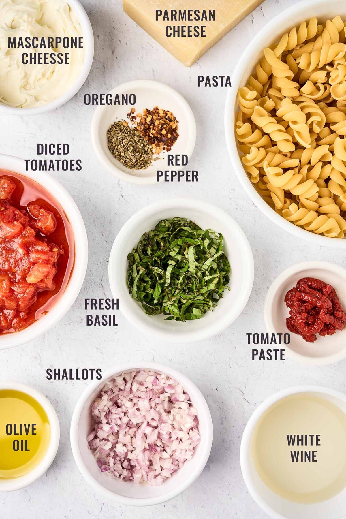 A labeled overhead view of the ingredients needed to make creamy tomato pasta with mascarpone; pasta, Parmesan cheese, mascarpone, diced tomatoes, fresh basil, shallots, white wine, tomato paste, and olive oil.