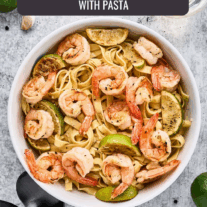 Garlic lime shrimp pasta garnished with lime wedges in a white bowl on a light stone surface, surrounded by garlic cloves, peppercorns, limes, and white wine.