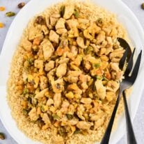 An oval serving platter of couscous topped with Persian style apricot chicken and pistachios with serving utensils, surrounded by a prep bowl of the spice blend, dried apricots, and chopped pistachios.