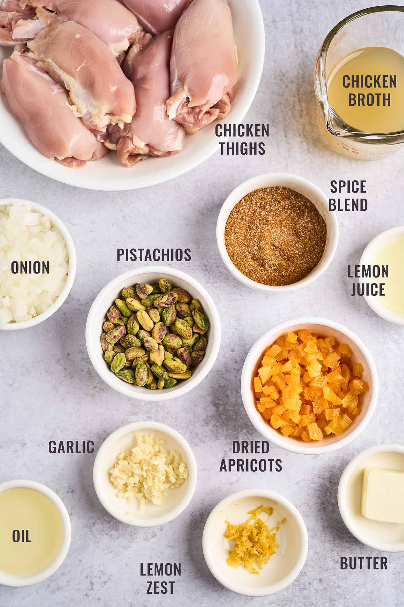 A labeled, overhead view of the ingredients for Persian apricot chicken; boneless, skinless chicken thighs, chicken broth, spice blend, lemon juice and zest, dried apricots, butter, garlic, oil, and chopped onion.