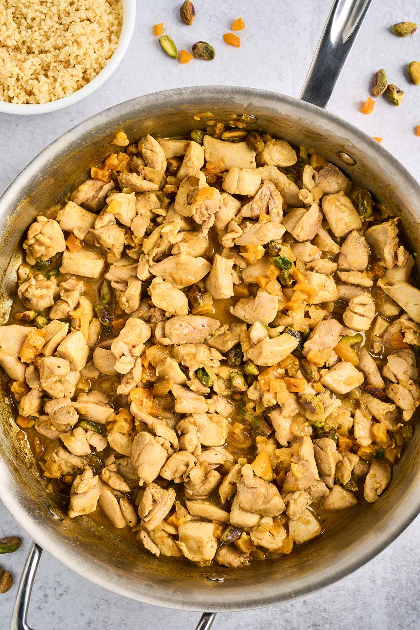 A frying pan filled with chunks of tender chicken thighs combined with spices, dried apricots, pistachio nuts, sautéed garlic and onion, lemon juice and zest, and butter, a bowl of couscous in the background.