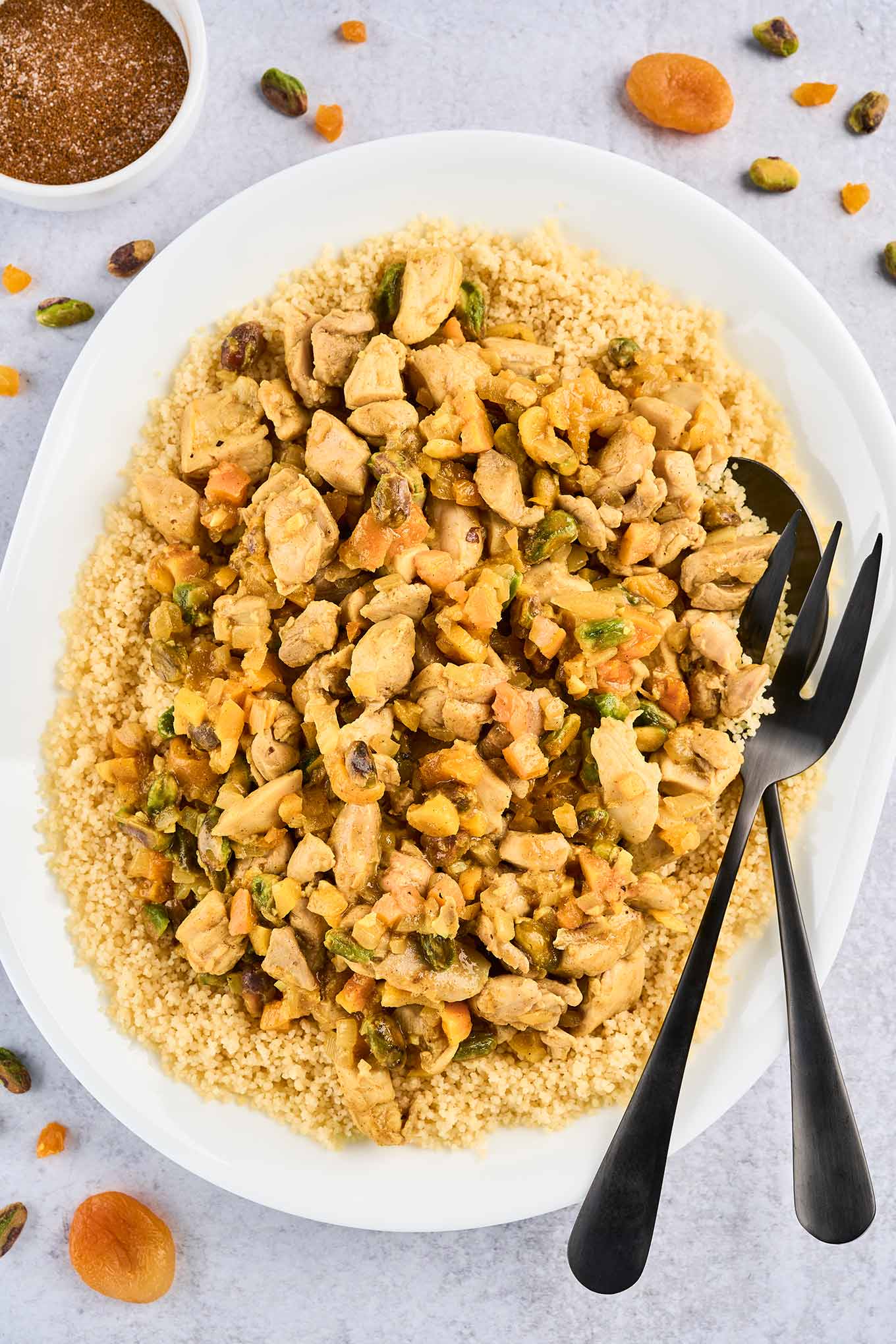 An oval serving platter of couscous topped with Persian style apricot chicken and pistachios with serving utensils, surrounded by a prep bowl of the spice blend, dried apricots, and chopped pistachios.