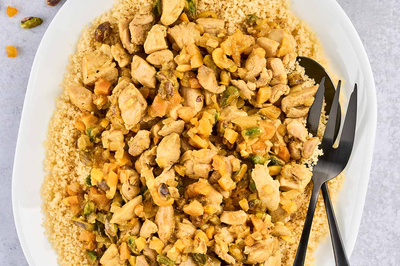 A close view of a platter of couscous topped with Persian-style apricot chicken, showing chopped pistachios, diced dried apricots, and lemon zest.