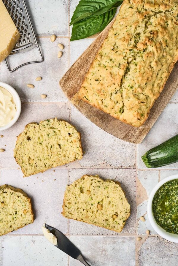 Three slices of savory pesto zucchini bread on a tile surface surrounded by a cutting board with the remainder of the loaf on it, a whole zucchini, a bowl of pesto, a dish of butter, Parmesan and a grater, pine nuts and basil leaves.