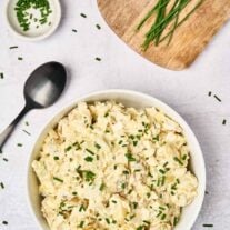 Sour cream potato salad in a white serving bowl, garnished with fresh chives, a black serving spoon to the left, a prep bowl of chopped chives, and fresh chives on a wooden board in the background.