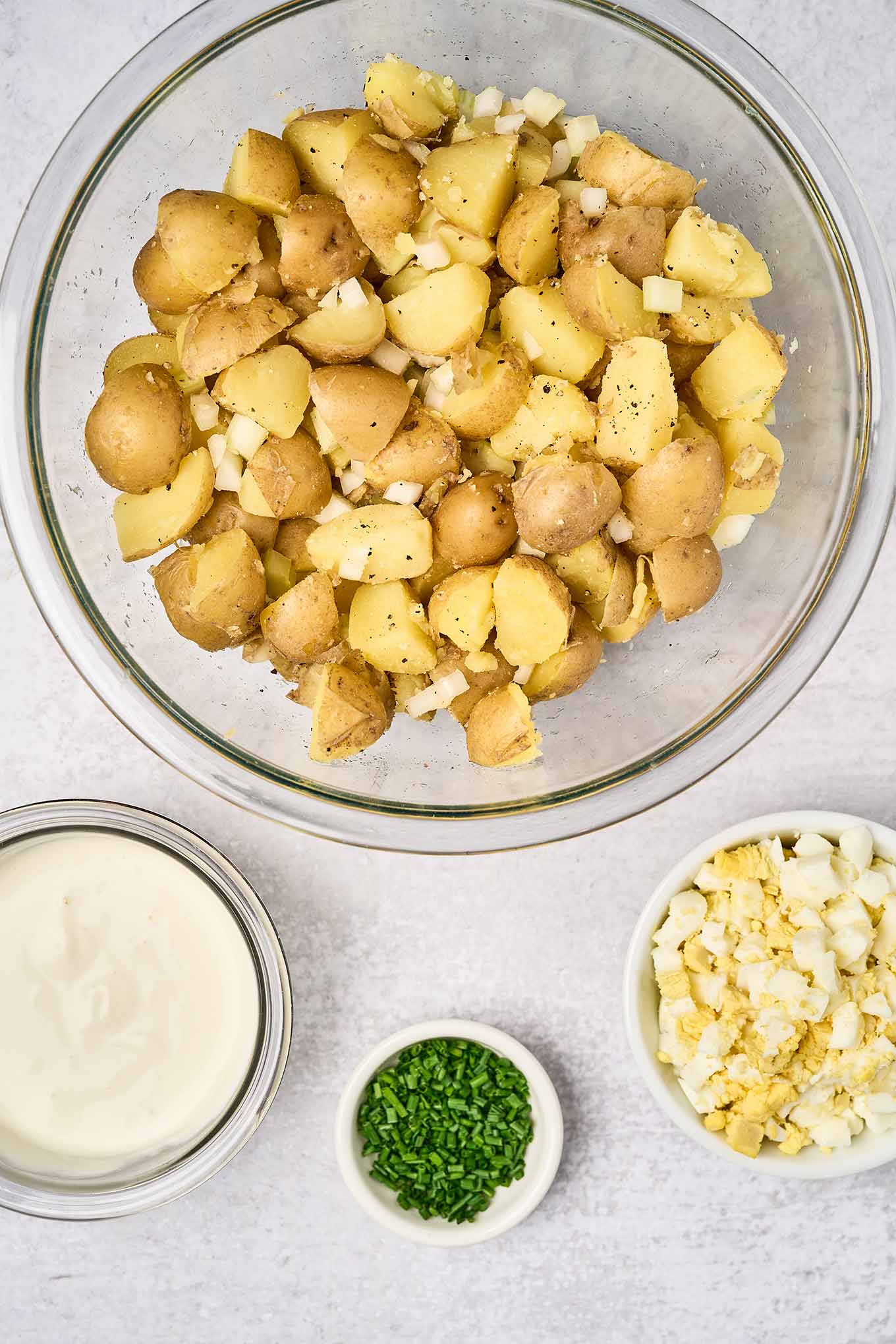 Boiled chunks of baby yellow potatoes and chopped celery and onion in a mixing bowl next to a small bowl of chopped hard boiled eggs, a prep bowl of chives, and a bowl of sour cream dressing - all ready to be combined.