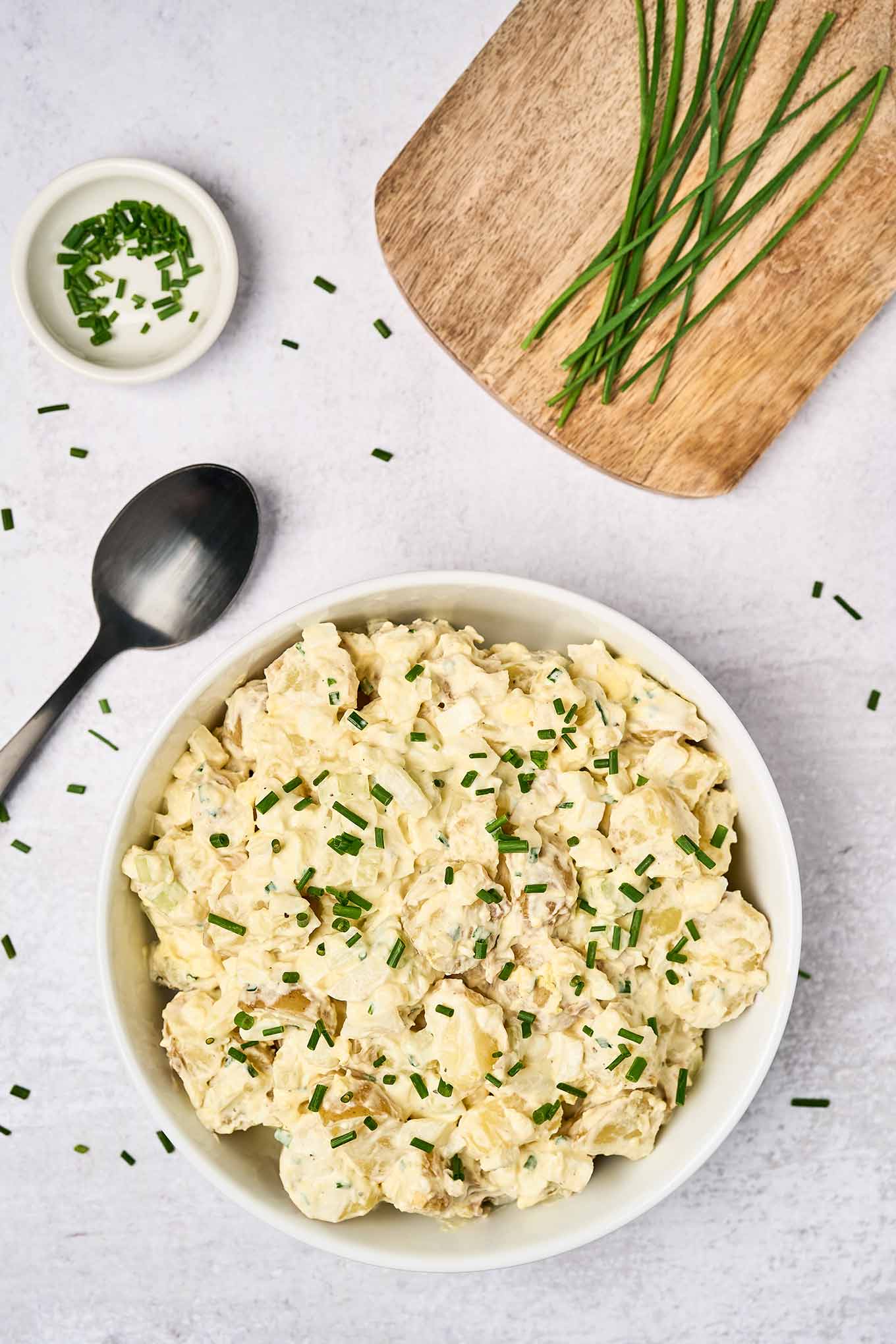 Sour cream potato salad in a white serving bowl, garnished with fresh chives, a black serving spoon to the left, a prep bowl of chopped chives, and fresh chives on a wooden board in the background.
