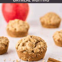 A single apple oatmeal muffin with more muffins and a whole apple in the background, cinnamon sticks and scattered oats in the foreground.