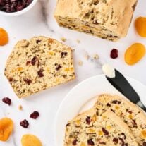 Sliced apricot bread with cranberries and walnuts on a white marble surface, surrounded by dried apricots, cranberries, and a buttered knife.